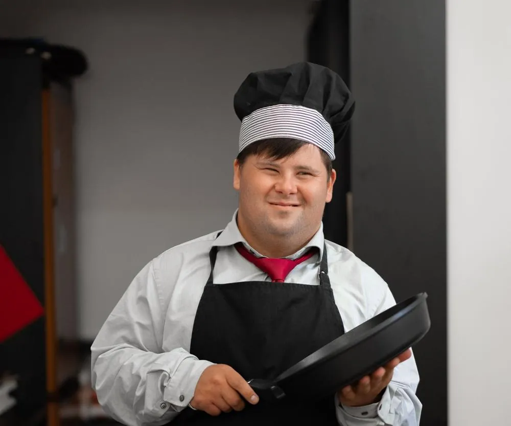 A smiling cook in an apron and chef’s hat holding a frying pan